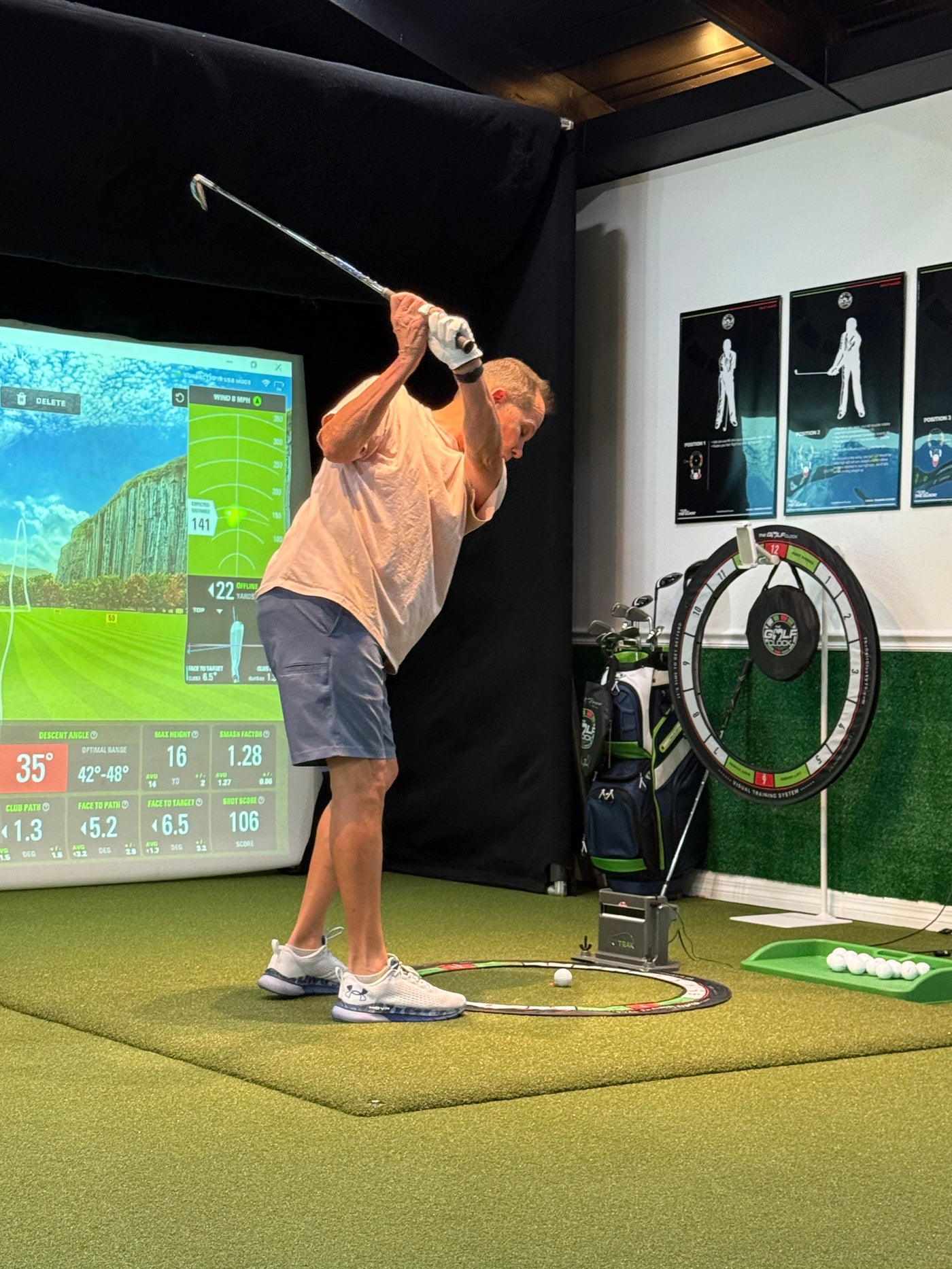 Golfer at the top of the backswing in an indoor simulator bay, launch monitor data on screen and The Golf Clock on the hitting mat